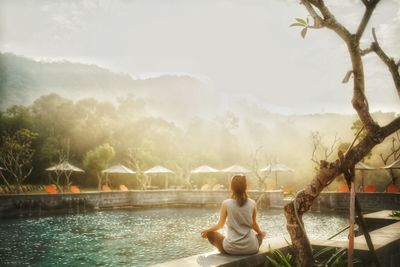 Rear view of woman sitting by lake against sky