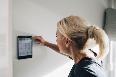 Side view of blond woman adjusting thermostat using digital tablet mounted on white wall at home