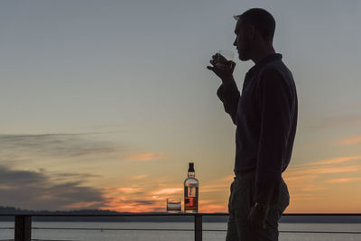 Side view of silhouette man having drink while standing by railing against sky