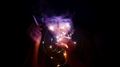 Close-up of man smoking while holding illuminated lighting equipment against black background