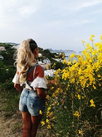 Rear view of woman standing by yellow flowering plants against sky