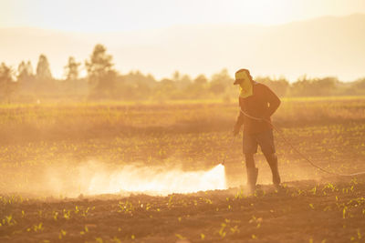 Full length of man spraying pesticides on field against sky during sunset