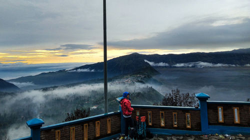 Man standing on railing by mountain against sky during sunset
