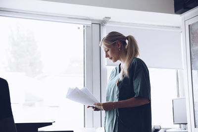 Female nurse reading documents while standing against window at doctor's office