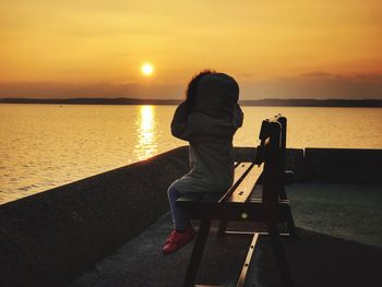 Rear view of woman sitting on sea against sky during sunset