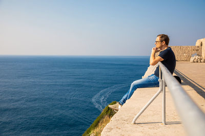 Side view of man sitting by sea against clear sky