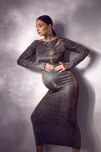 Trendy stylish beautiful pregnant woman in a tight silver sequined dress against a white wall 