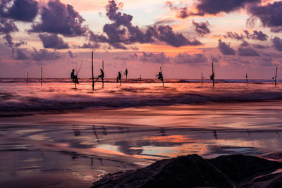 Group of distant anonymous workers sitting on poles with fishing rods while catching fish in waving sea in evening time