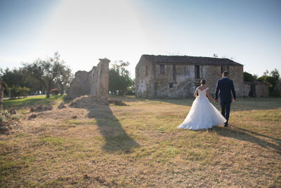 Full length rear view of newlywed couple walking by field by abandoned house against clear sky