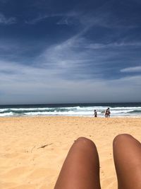 Cropped legs of woman relaxing at beach against blue sky