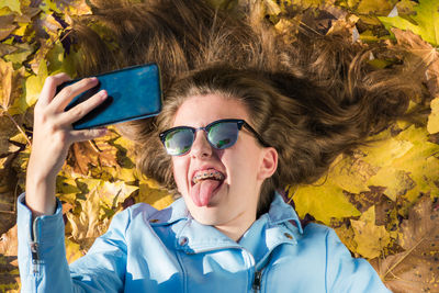 Teenager girl wearing sunglasses and lying down on the floor covered by yellow leaves while taking a photo with her mobile phone to herself. the girl is smiling and showing her tongue to the