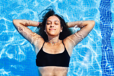 From above of charming young female in bikini swimming in pool with clear water while looking at camera in summer