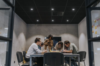 Multiracial male and female colleagues writing while discussing in meeting at illuminated board room