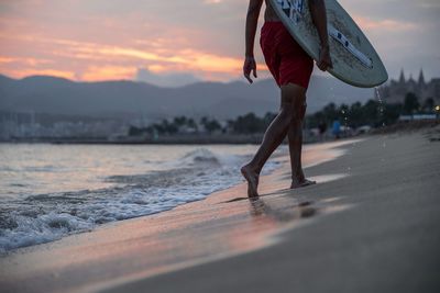 Low section of man carrying surfboard at beach during sunset