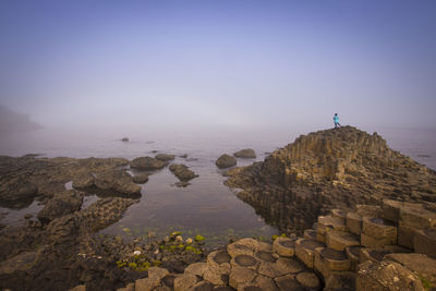 Mid distance view of woman standing on rocks by sea against sky during foggy weather