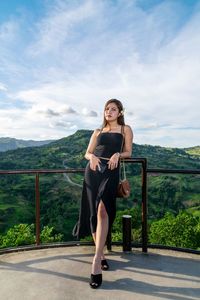 Full length portrait of young woman standing on railing against sky