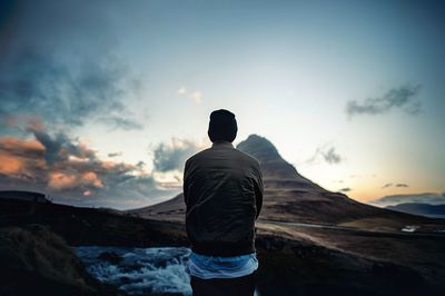 Rear view of man standing on shore against sky during sunset