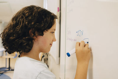 Side view of boy with curly hair writing on whiteboard at classroom