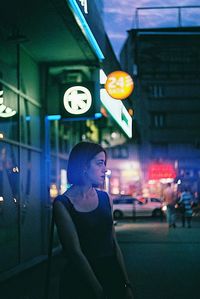 Woman standing on sidewalk against illuminated building in city at night