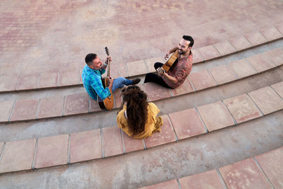 Full body of content male musicians playing songs on acoustic guitars and looking at each other while sitting on steps with cheerful woman on street