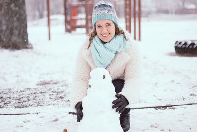 Portrait of smiling young woman making snowman at park during winter