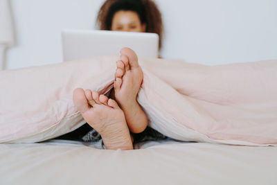 Young ethnic barefoot female covered with blanket sitting in bed and browsing laptop at home