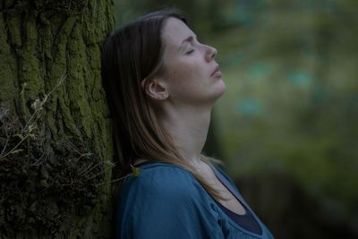 Side view of young woman against tree trunk