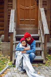 Father and son in red hats and knitted scarves sitting on porch stairs. wooden house logs in autumn