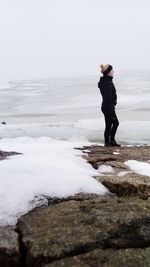 Side view of woman standing on rock formation by frozen lake against sky during winter