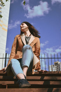 Young woman looking away while sitting on railing against sky