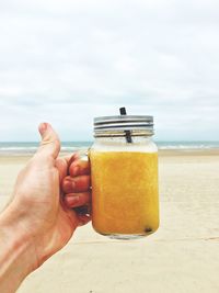 Cropped image of hand holding drink in mason jar at beach against sky