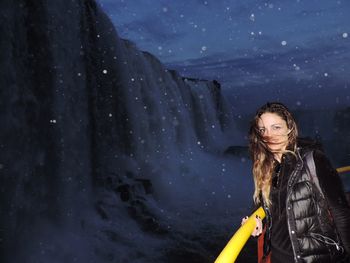 Portrait of woman standing against waterfall at dusk during snowfall