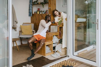 Smiling woman sharing book with man sitting on seat at home seen through doorway