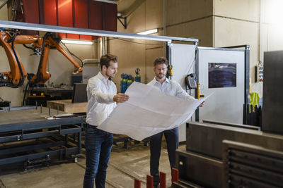 Businessmen studying blueprint of business while standing against automated machine at factory