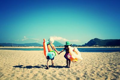 Rear view of siblings walking with inflatable rings on beach against sky