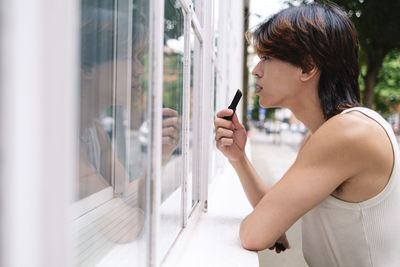 Young gay man applying lipstick looking at window