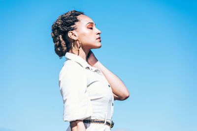Trendy ethnic female model in casual white dress with traditional african accessories with closed eyes while standing in grassy meadow near farm building