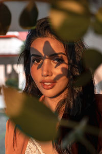 Charming young ethnic female with long dark hair looking at camera while standing behind green leaves of tropical plant