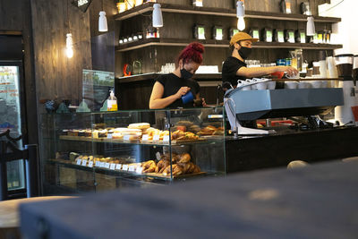 Busy baristas in protective masks making delicious coffee while standing at counter in cozy coffee house during coronavirus pandemic