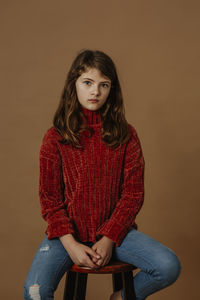 Portrait of serious girl sitting on stool against brown background