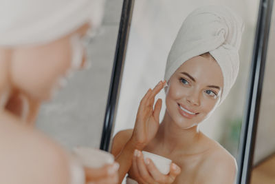 Smiling young woman applying cream on face while looking in mirror