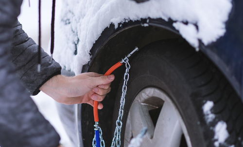 Cropped hand of man holding chain equipment by car tire during winter