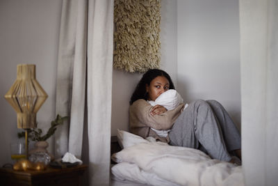 Pensive young woman sitting on bed and hugging pillow