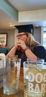 Woman wearing mortarboard sitting by drink glasses at restaurant