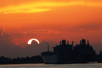 Silhouette factory by sea against sky during sunset