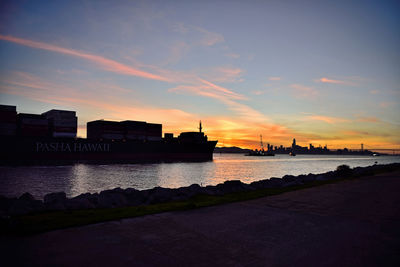 Silhouette buildings by river against sky during sunset