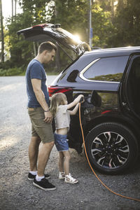 Portrait of mother, father and two daughters standing by car at electric vehicle charging station