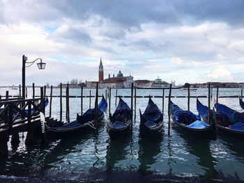 Gondolas moored in canal against cloudy sky