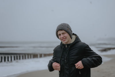 Portrait of a teenager at the sea, a guy in a winter hat and a warm jacket squints against the stron