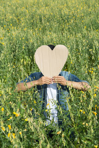 View of man covering face with heart shape while standing in field